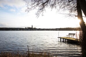 Un lago tranquillo con un pontile e una panchina, circondato da alberi e un edificio in lontananza.
