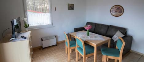 Ein Zimmer mit Schreibtisch aus Holz, einem Tisch mit Stühlen, einer Couch, einem Heizkörper, einem Fenster und einem Deckenventilator.