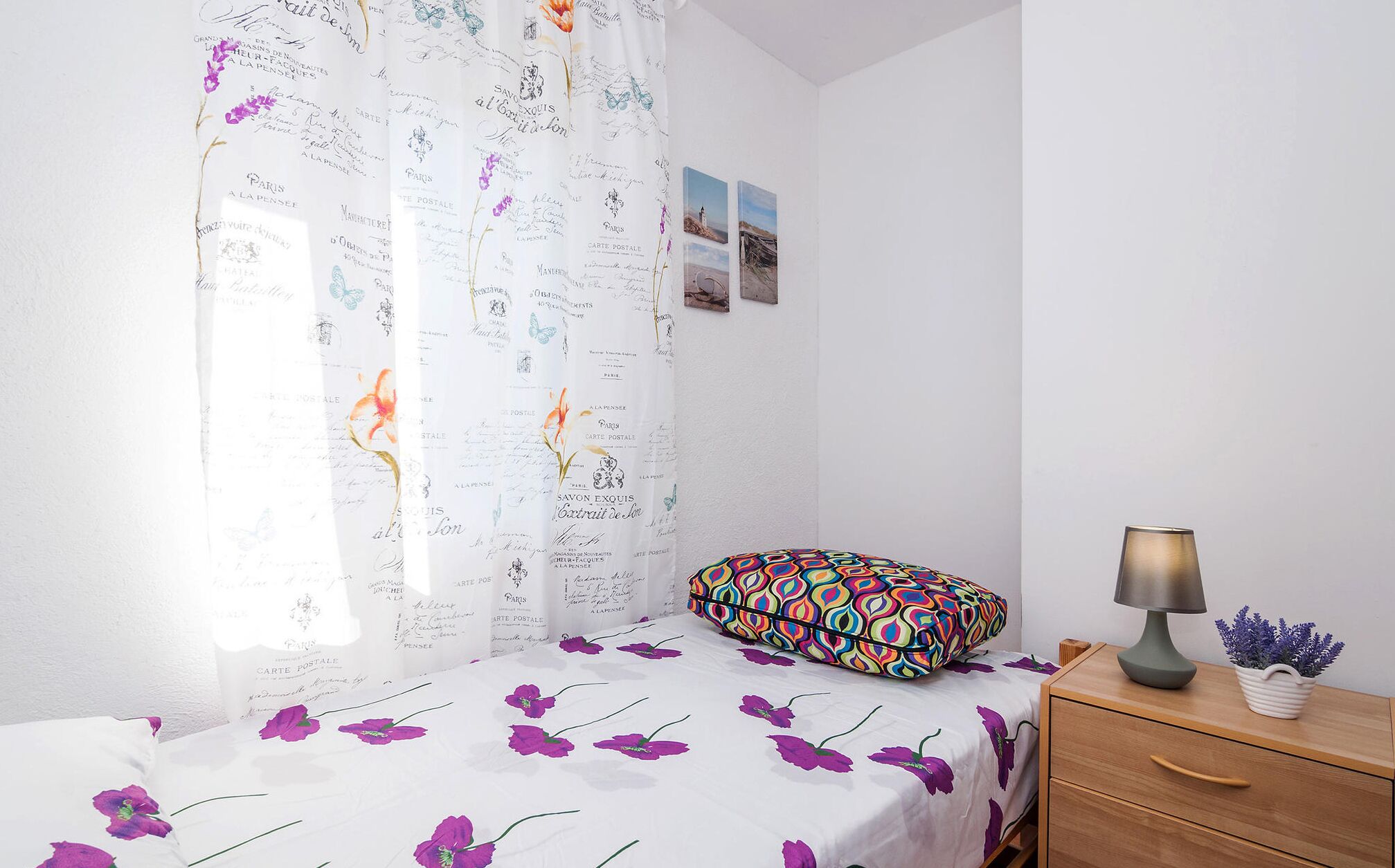 A bedroom with a bed covered in a floral-patterned bedspread, a colorful pillow, a nightstand with a lamp, and a wall with framed pictures.