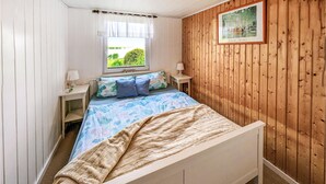Un dormitorio con cama, ventana con cortinas, mesita de noche, lámpara y pared de madera con un cuadro enmarcado.