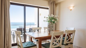 Zona pranzo con tavolo in legno e sedie bianche con cuscini verdi, un vaso con fiori bianchi e vista mare attraverso ampie vetrate.