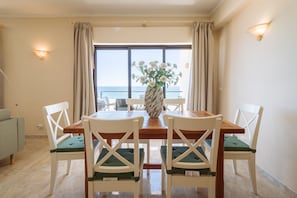 A dining area with a table and chairs, a vase with flowers, and a view of the sea through a large window.