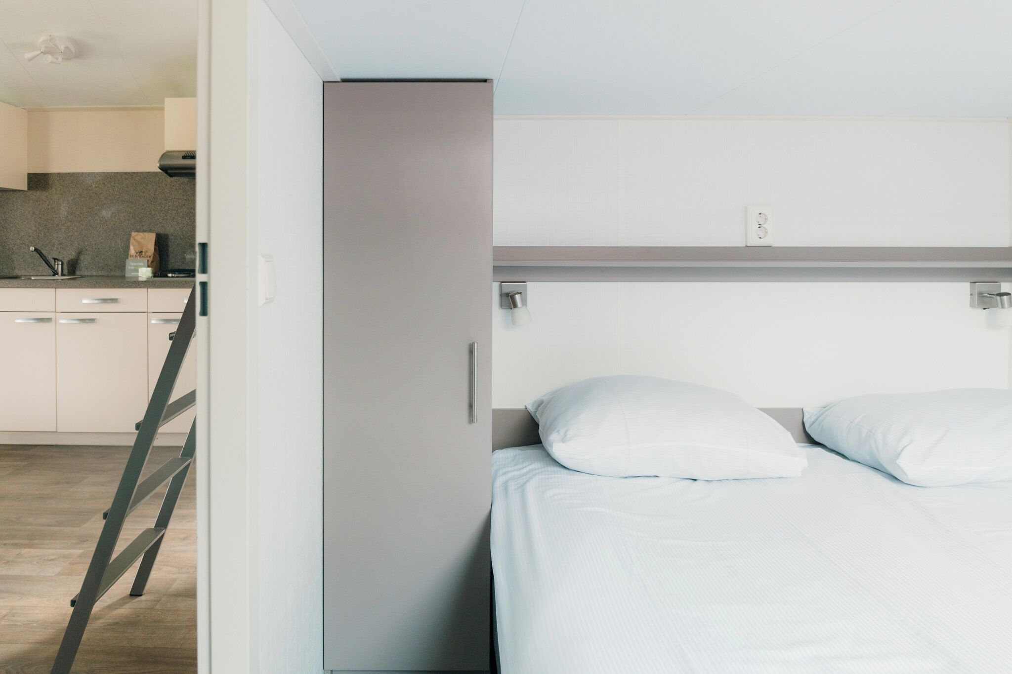 Un dormitorio moderno con cama, almohadas blancas, un armario y una kitchenette al fondo.