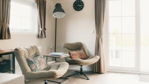 Ein Wohnzimmer mit zwei Sesseln, einem Beistelltisch, einer Lampe, einer Uhr und einem großen Fenster mit Vorhängen.