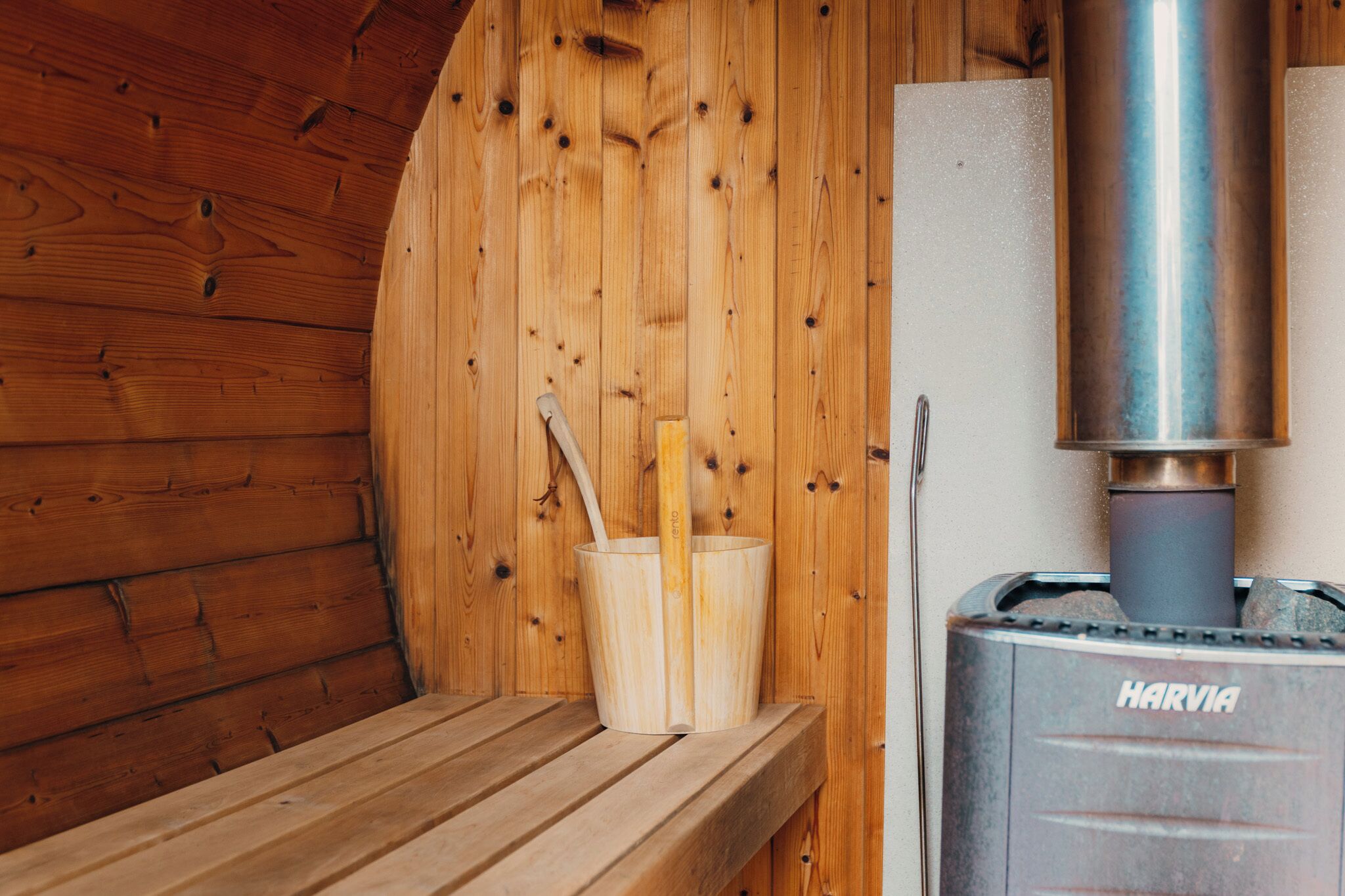 A wooden sauna room with a Harvia sauna heater, a wooden bench, and a bucket with wooden sticks.