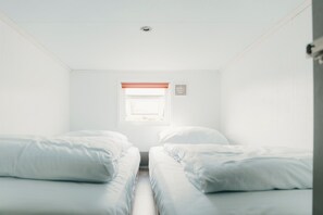 Een kleine, witte kamer met twee bedden, een raam met zonwering en een ventilatie rooster.