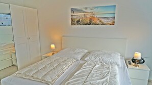 A neatly made bed with white bedding and two bedside tables, one with a lamp and the other with a radio.