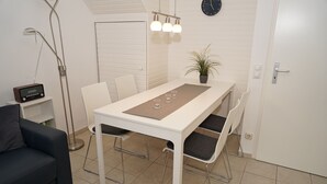A modern, well-lit room with a dining table, chairs, a potted plant, a clock, and a radio on a shelf.