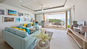 Ein modernes Wohnzimmer mit einem hellblauen Sofa, gelb-blauen Kissen, einem Couchtisch mit Blumenvase und einem großen Fenster mit Blick auf einen Balkon und Gartenmöbel.