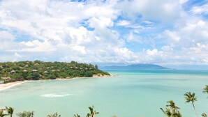Ein tropischer Strand mit klarem türkisblauem Wasser, ein üppiger, grüner Hügel mit Gebäuden und ein teilweise bewölkter Himmel.
