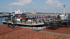 Un bateau amarré, doté d’un espace de repos couvert avec des tables, d’un vélo, et offrant une vue sur le port de plaisance et l’eau.