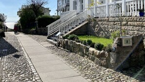 Un edificio bianco con una scala in pietra che conduce a un balcone, un vialetto in sampietrini e un cielo sereno.