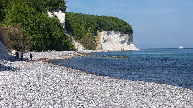 Una spiaggia di ciottoli con una scogliera bianca e vegetazione, una barca in acqua e persone che passeggiano.