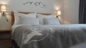 Una cama bien tendida con un edredón gris con estampado floral blanco, flanqueada por almohadones a cuadros. Dos lámparas de pared proporcionan iluminación, y se observa una decoración de aves sobre la cama.