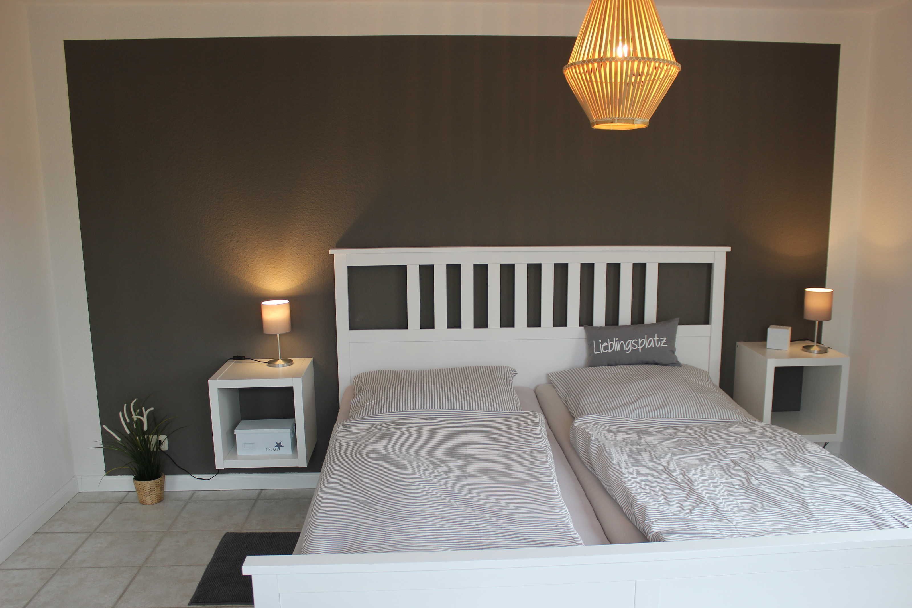 Een slaapkamer met een wit bed, twee nachtkastjes met lampen, een potplant en een donkere muur.