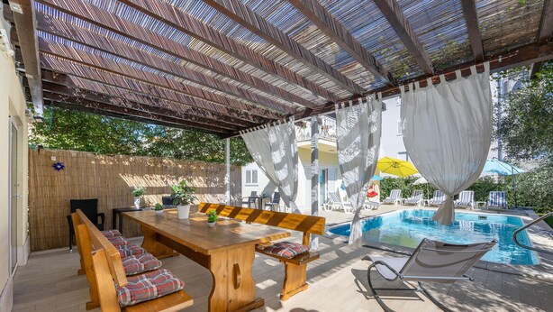 An outdoor pool area with a wooden table and chairs, a covered seating area, and a pool with a yellow umbrella. - Beautiful home in Kastel Stafilic with sauna (Kastel Stafilic)