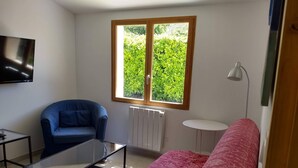 Ein Wohnzimmer mit blauem Sofa, rotem Sofa, Glastisch, blauem Sessel, weißem Heizkörper und einem Fenster mit Blick auf Grünflächen.