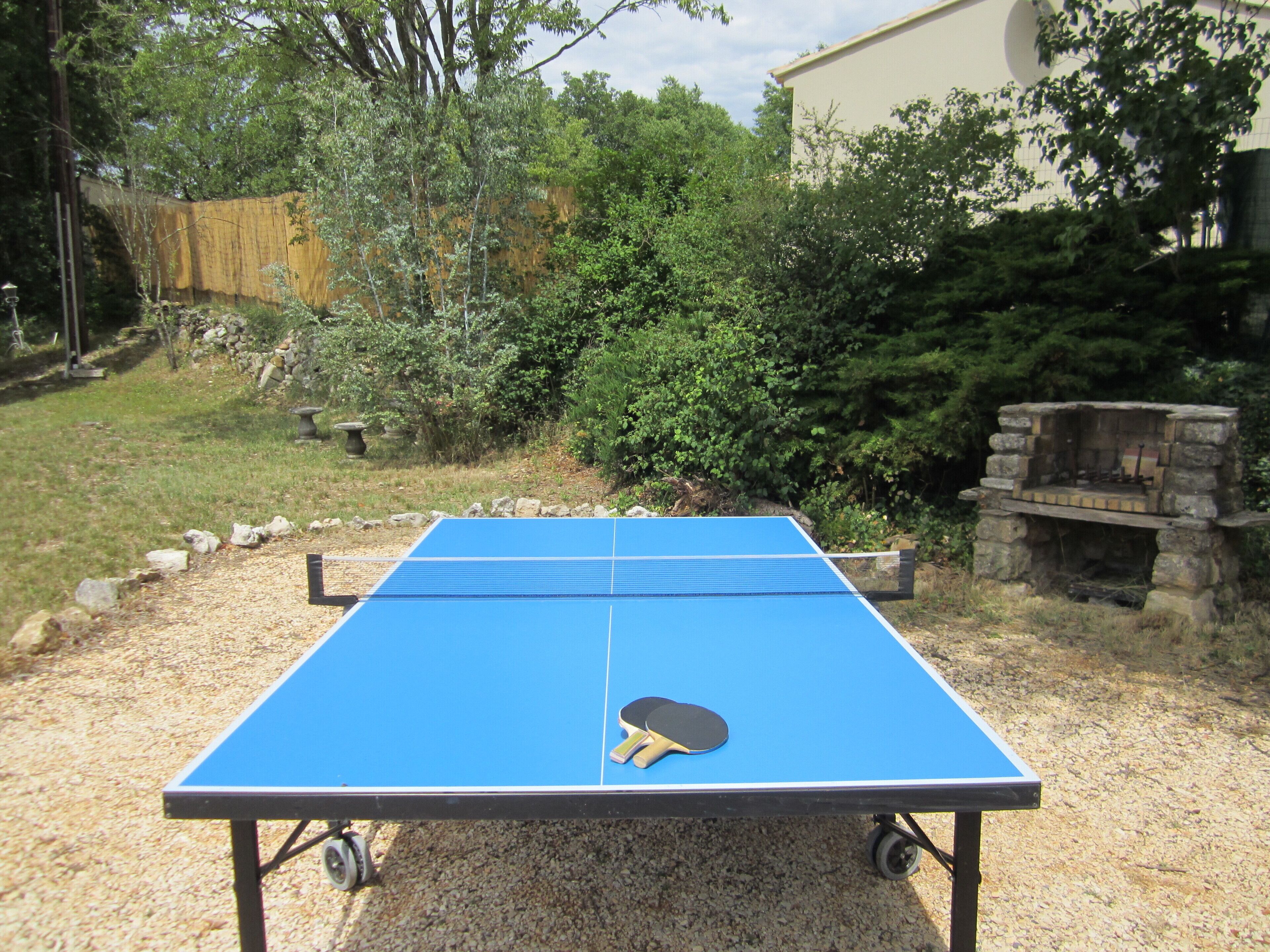 A ping pong table with a paddle in the center, surrounded by a gravel area with a stone bench and a stone fireplace in the background.