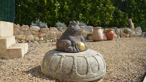 Eine Steinfroschstatue auf einem dekorativen Sockel in einem Garten, im Hintergrund ein Hecken und eine Steinmauer.