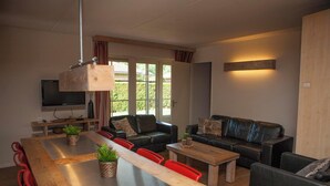 A modern living room with a wooden dining table, red chairs, a black leather sofa, and a television on a wooden stand. - Wilbrink 6 to 8-person bungalow (Voorthuizen)