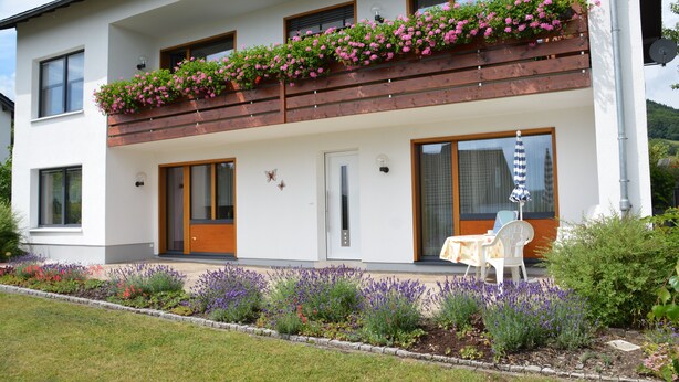 Una casa moderna de dos pisos con un balcón decorado con flores rosadas, una puerta principal y un pequeño patio con mesa y sillas.