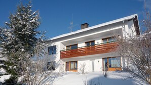 Un edificio moderno de dos pisos con un balcón, rodeado de árboles y arbustos cubiertos de nieve.