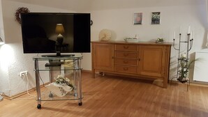 Een woonkamer met een flatscreen televisie op een glazen voetstuk, houten kasten, een klein tafeltje met een lamp en een kamerplant.