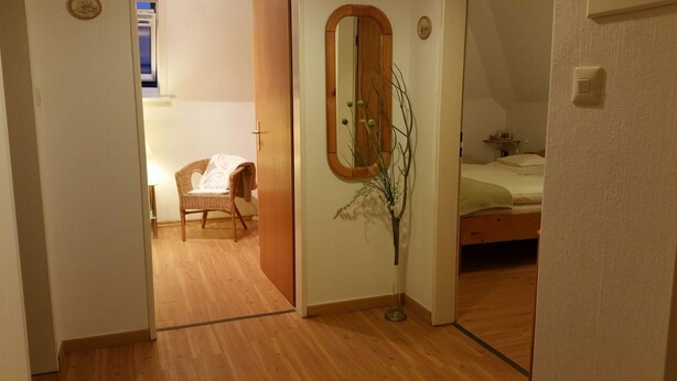 Een kamer met een houten vloer, een bed met een groene deken, een stoel met een witte deken en een spiegel met een bloevas.