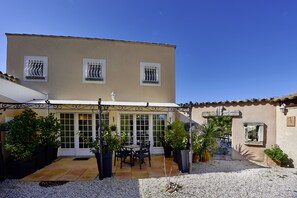 Un edificio a due piani con un’area patio dotata di tavolo e sedie, circondata da piante in vaso e un vialetto ghiaioso.