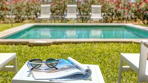 Zona de piscina con piscina, sillas de descanso blancas y una mesa con un par de gafas y una revista enrollada.