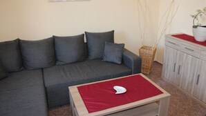 Um sofá cinzento com almofadas, uma pequena mesa de madeira com um tecido vermelho, um vaso branco com ramos e um armário de cor clara.