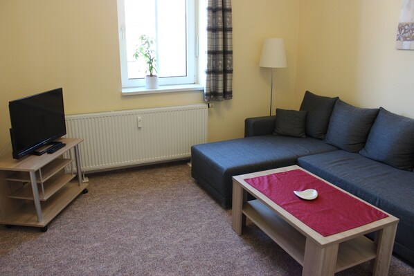 A living room with a sofa, a TV on a stand, a table with a red cloth, and a window with curtains.