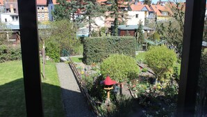 Vista para um jardim com um caminho, uma construção com telhado vermelho e diversas plantas.