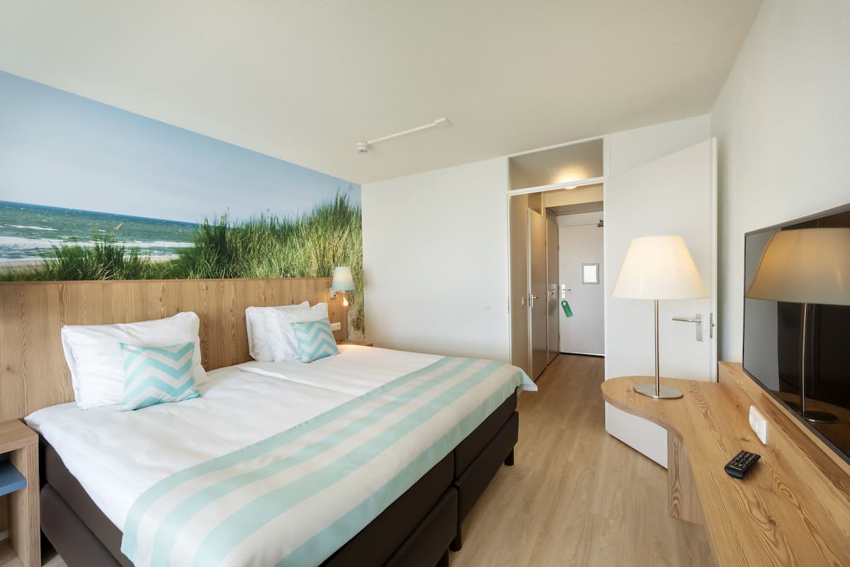Un dormitorio con cama, televisión, lámpara y vista a la playa.