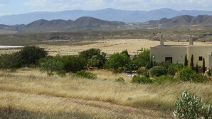 A desert landscape with mountains in the background and a building with a flat roof and chimneys in the foreground.