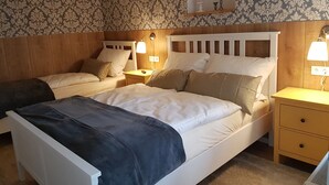 A bedroom with two beds, wooden headboards, a nightstand with a lamp, a small shelf with decorative items, and patterned wallpaper. - Apartment Rose       (Obermöllrich)