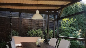 A covered patio with a wooden table and white cushioned chairs, surrounded by a wooden fence and greenery. - Apartment Rose       (Obermöllrich)