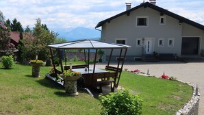 A gazebo with a table and chairs in a garden area, a large house with a driveway, and mountains in the background. - Holiday house on the outskirts of Klagenfurt with a wonderful view (Klagenfurt am Wörthersee)