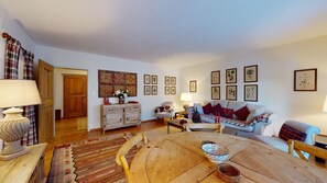 A cozy living room with a wooden coffee table, a sofa, and a chair. There are framed pictures on the wall and a wooden cabinet.