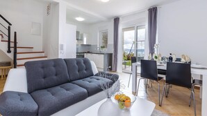 Una sala de estar moderna con un sofá gris, un comedor con una mesa blanca y sillas negras, una cocina al fondo y un balcón con vistas.