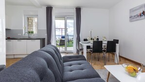 Una sala de estar moderna con un sofá gris, un comedor con mesa y sillas blancas, y una cocina con armarios blancos y ventana con vistas a un jardín.