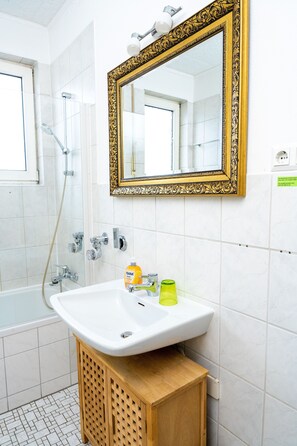 Un baño con un lavabo blanco, un mueble de madera, un espejo con un marco decorativo y una zona de ducha.