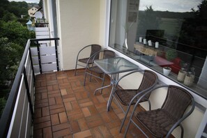 Un balcón con piso de baldosas, sillas de metal, una mesa de vidrio y vistas a zonas verdes y edificios.