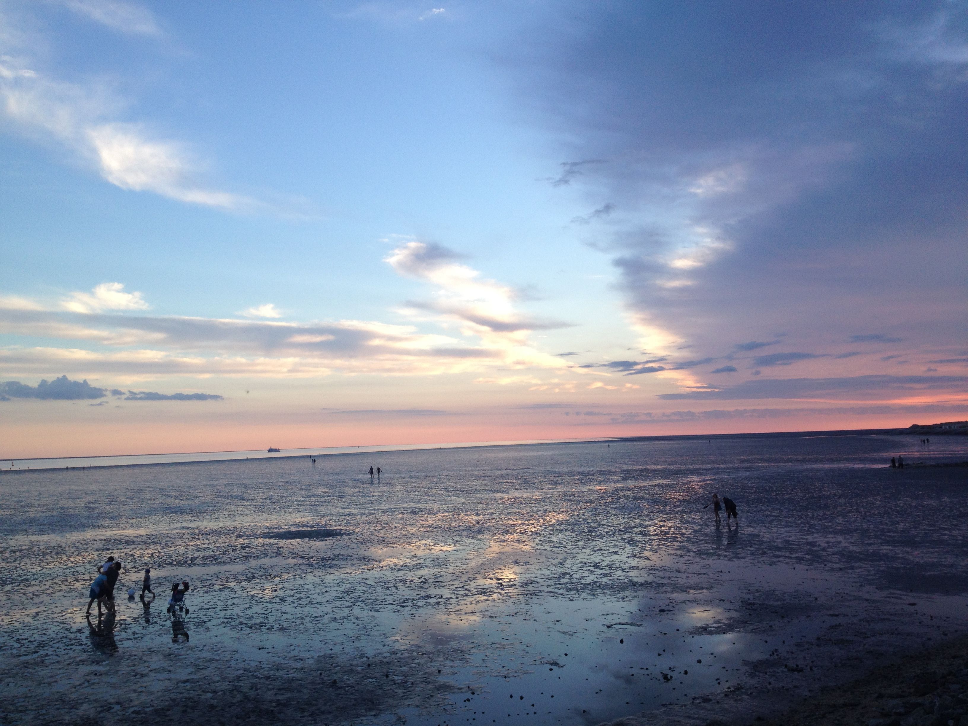 A beach with people playing and walking, and a distant ship on the horizon.