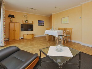 A living room with a black leather sofa, a glass coffee table, a dining area with a table and chairs, a TV mounted on the wall, and a wooden cabinet. - N26 (BC.a5) - Stranddomicil Steiner (Büsum)