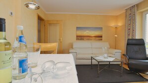 Une chambre d’hôtel avec un plancher en bois, un canapé blanc, un fauteuil en cuir noir, une table basse en verre et un tableau représentant un coucher de soleil accroché au mur.