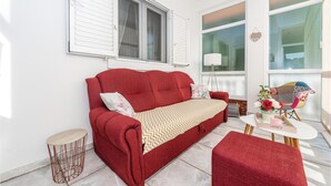 Une pièce chaleureuse avec un canapé rouge, une petite table ronde et une chaise. Il y a deux fenêtres avec des volets blancs, et une lampe sur la table.
