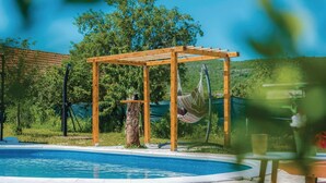 Un espace piscine agrémenté d’une pergola en bois, d’un hamac et d’un tronc d’arbre.