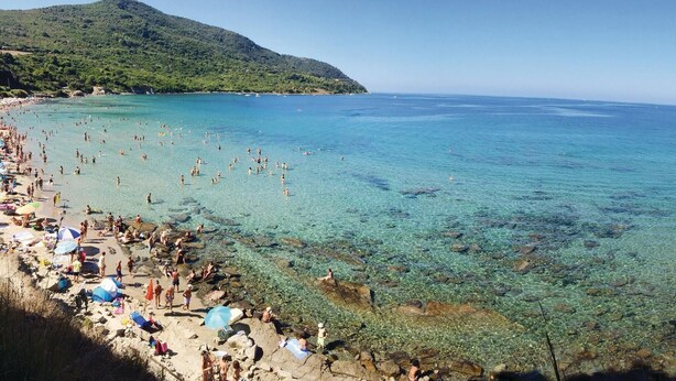 A beach with clear shallow waters, a sandy shore, and a mountain in the background.