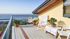 A balcony with tiled flooring, white chairs, potted plants, and a view of the sea and cityscape.